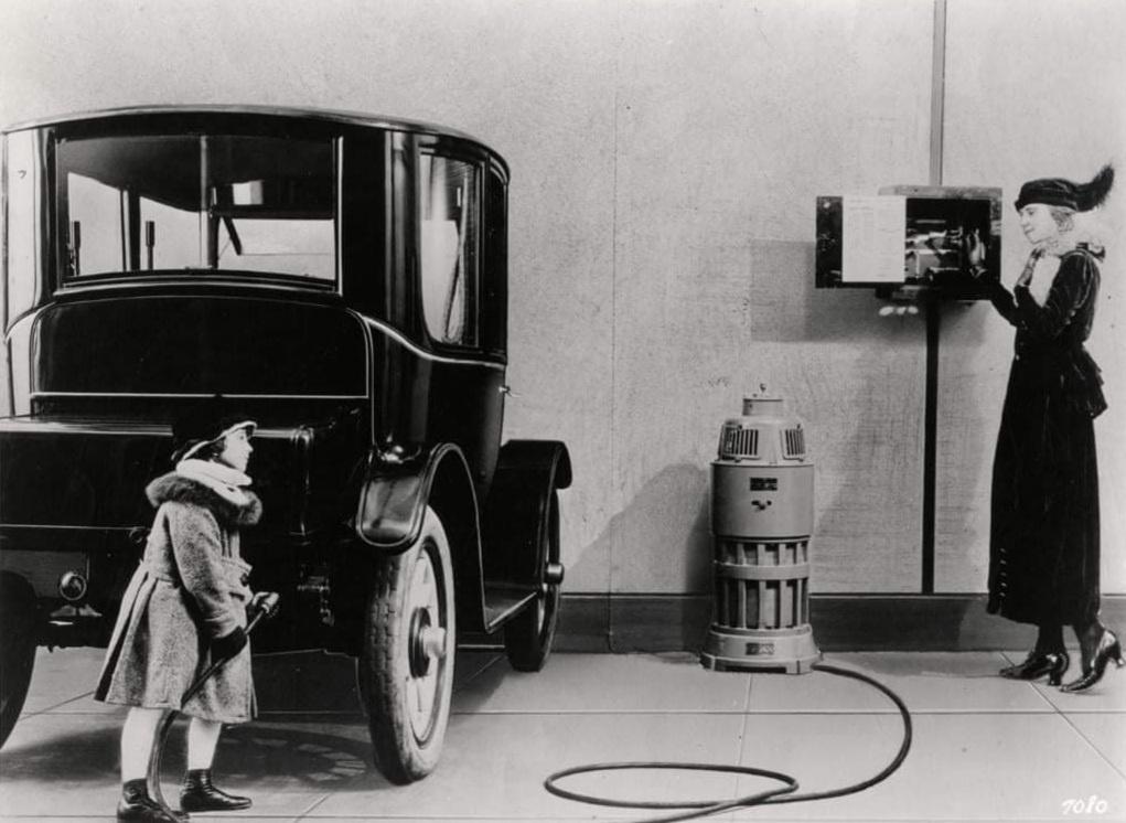 Leer of a girl charging a Baker electric automobile, whereas a woman stands subsequent to a wall-mounted electrical box in background. c.1914.